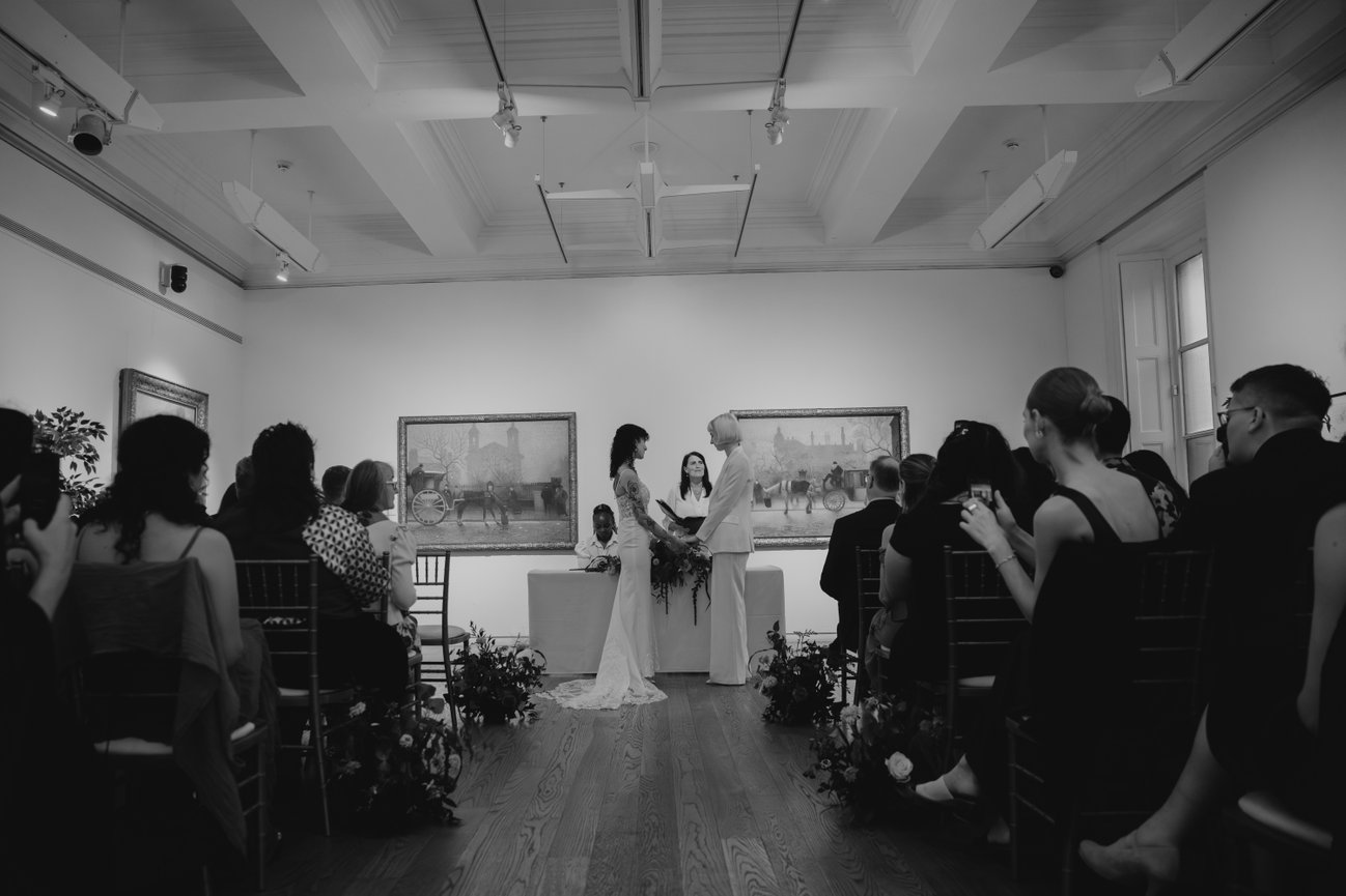 Wide editorial shot from the back of the room during a ceremony at Manchester Art Gallery, showing seated guests in a classic black and white aesthetic.