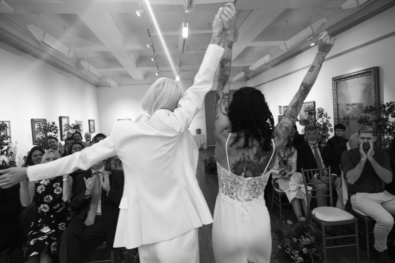 Editorial wedding photography of a same-sex couple celebrating their ceremony at Manchester Art Gallery, surrounded by cheering guests in a grand, art-filled hall.