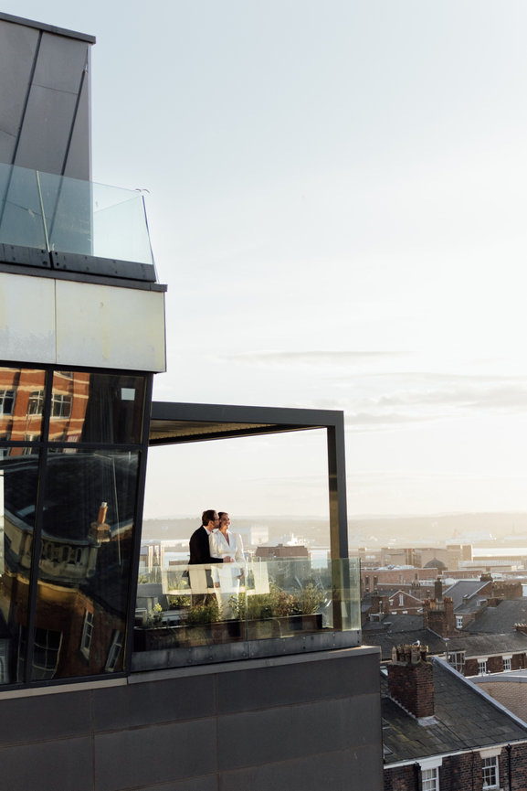 Modern editorial wedding photography of a bride and groom on a balcony at Hope Street Hotel, silhouetted against the Liverpool cityscape at sunset.