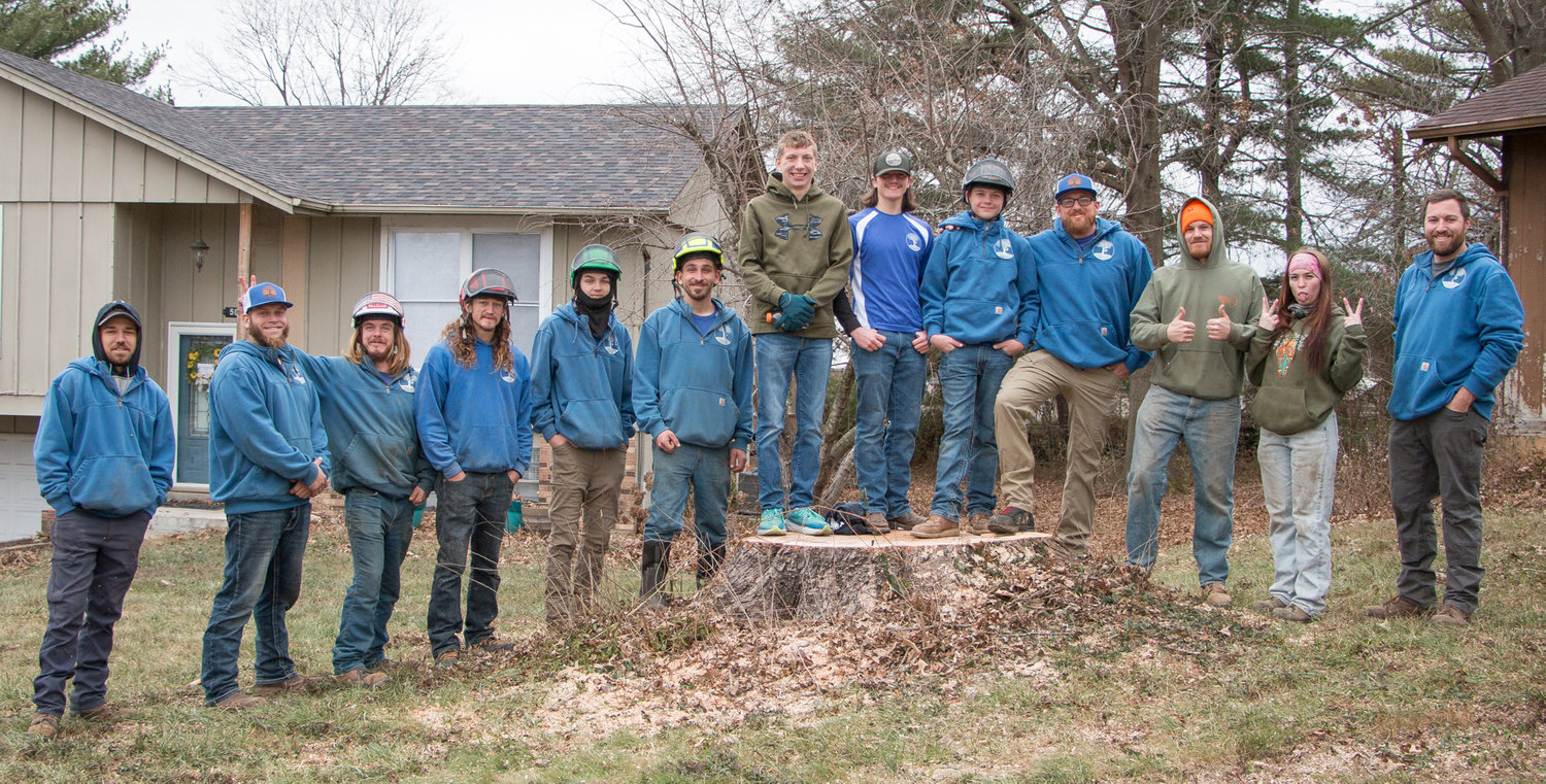 A group of tree trimmers stand around a large tree stump in a yard.