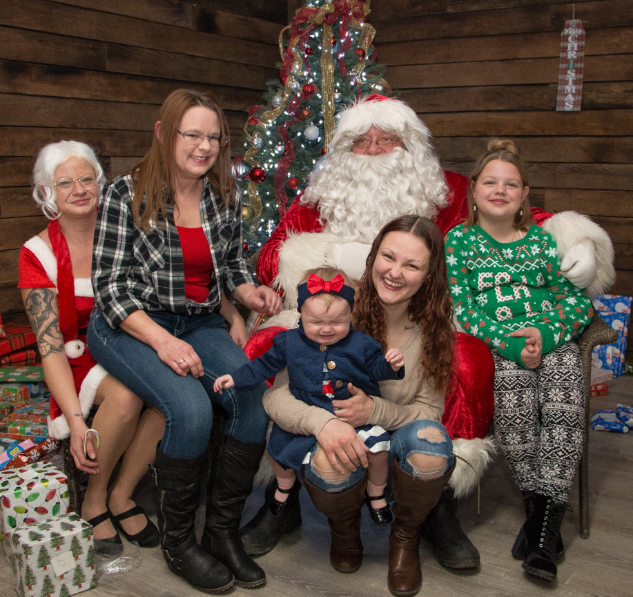 Family poses with Santa and Mrs. Claus by a Christmas tree, surrounded by gifts, with festive attire.