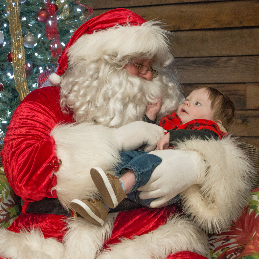 Santa Claus holding a baby in his arms, sitting in front of a decorated Christmas tree.