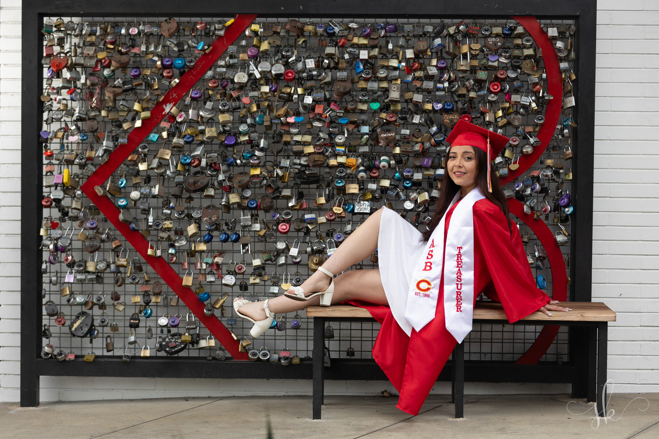 Young woman in red graduation gown sitting on a bench in front of a lock wall