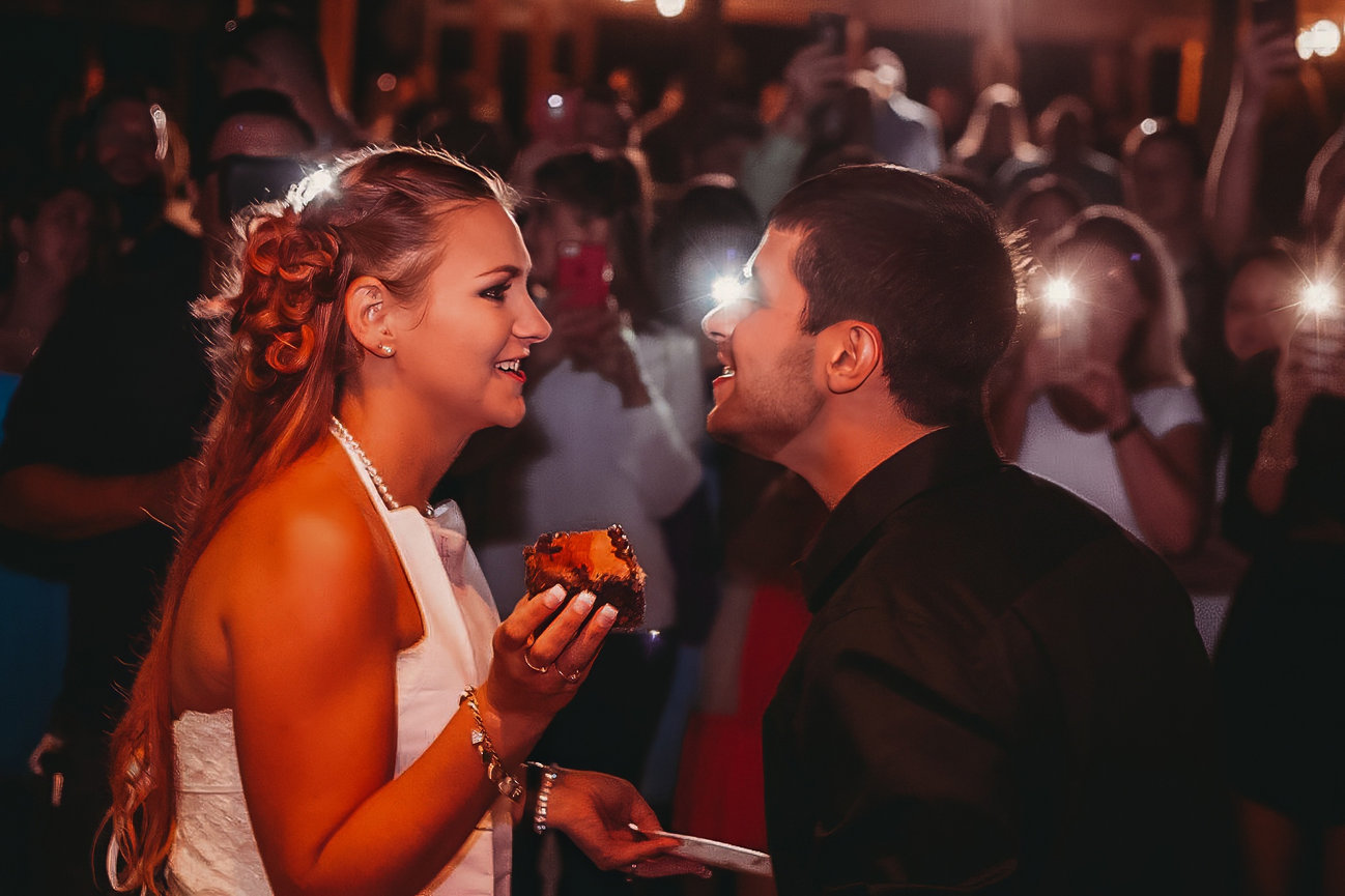 A couple smiling at each other during a wedding reception, with the bride holding a piece of cake.
