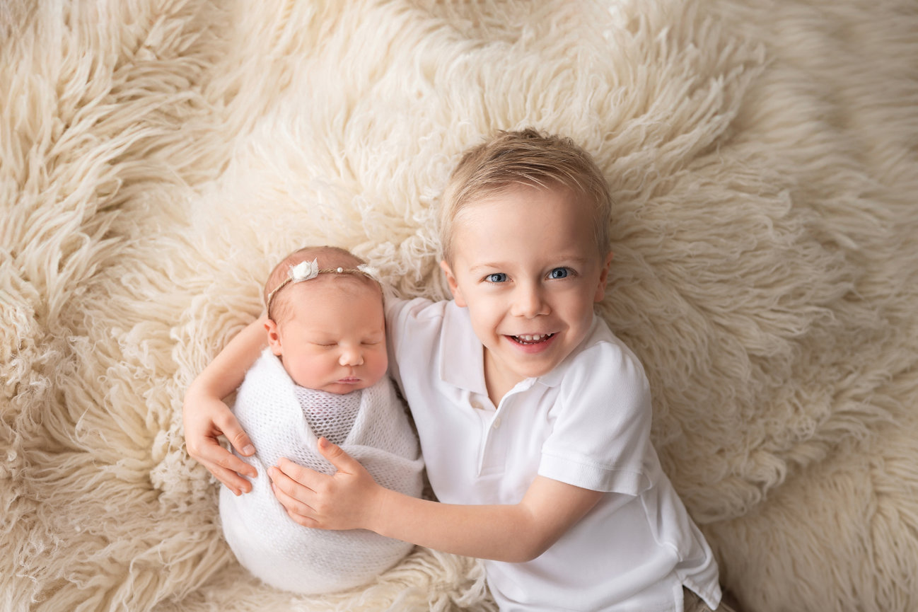 Big brother snuggling newborn sibling on white fur rug during studio newborn session in Manasquan, NJ