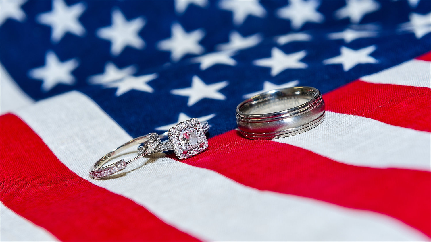 Close-up of a diamond engagement ring and a wedding band placed on an American flag, with the rings positioned on the red and white stripes against a blurred blue field with white stars.
