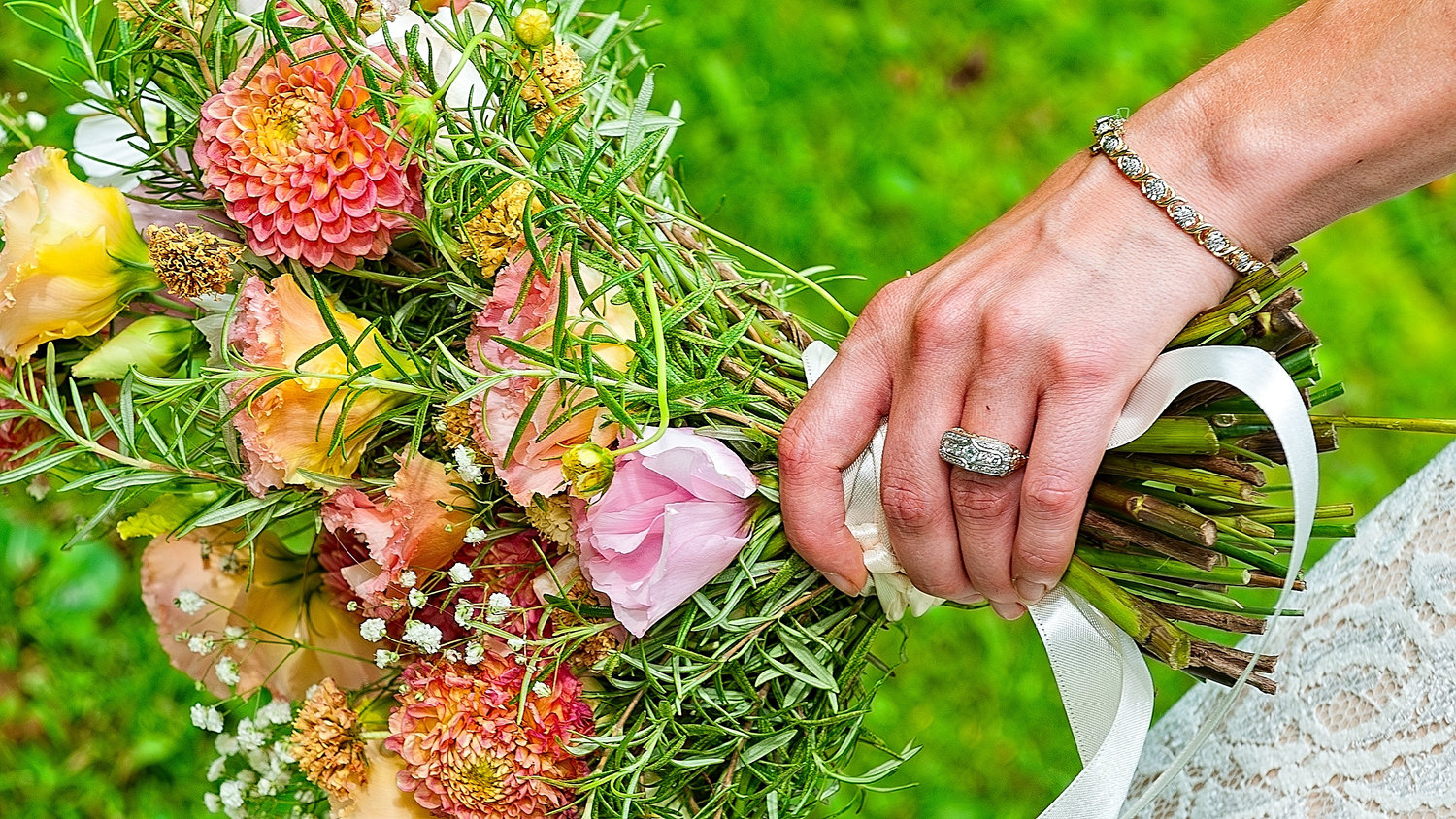 Close-up of a bride’s hand holding a bouquet of colorful flowers with greenery, wearing a vintage-style diamond ring and matching diamond bracelet, with a green grass background