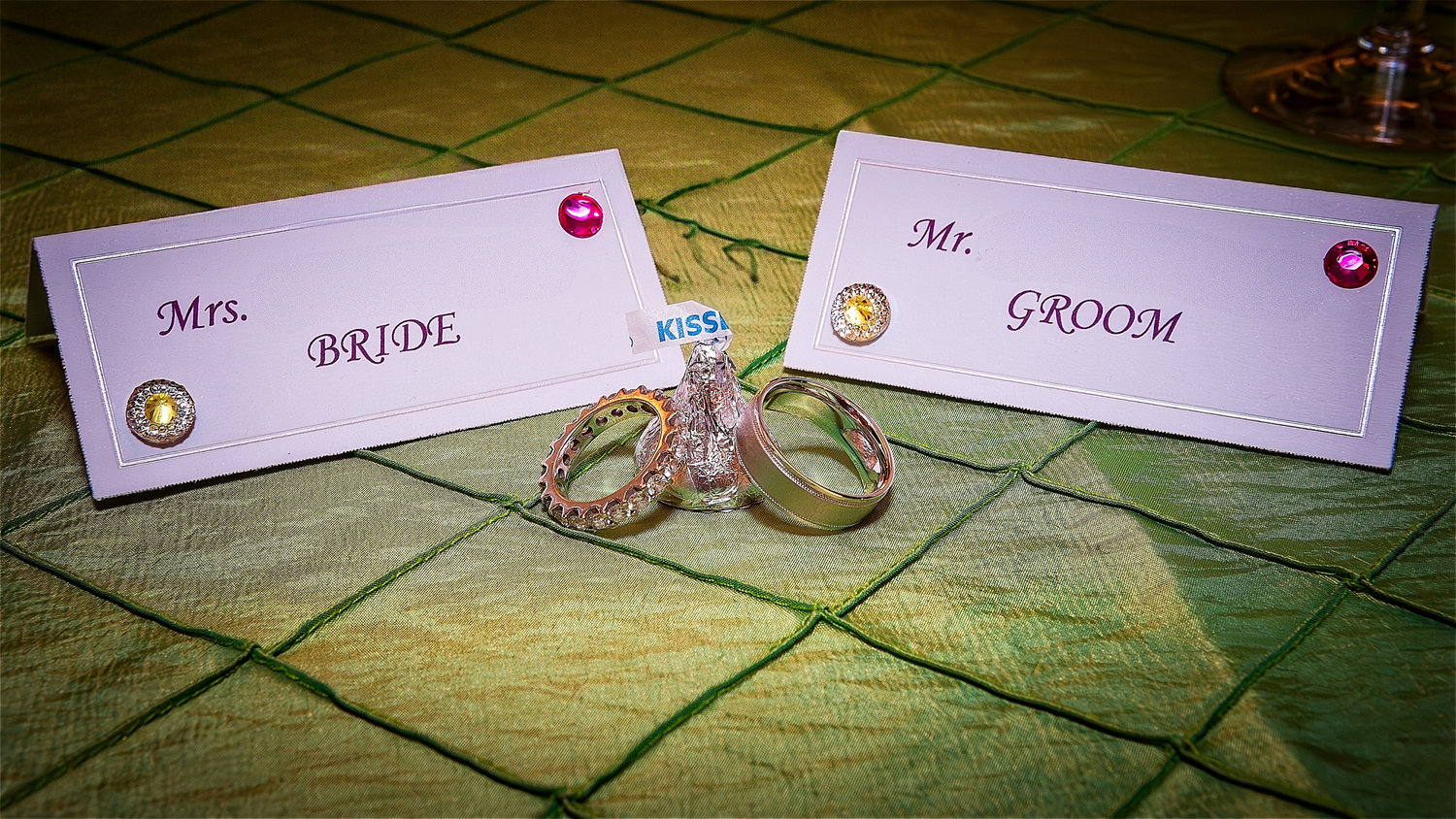 Wedding place cards labeled “Mrs. Bride” and “Mr. Groom” with jeweled accents, displayed on a green textured tablecloth alongside a Hershey’s Kiss and two wedding rings.