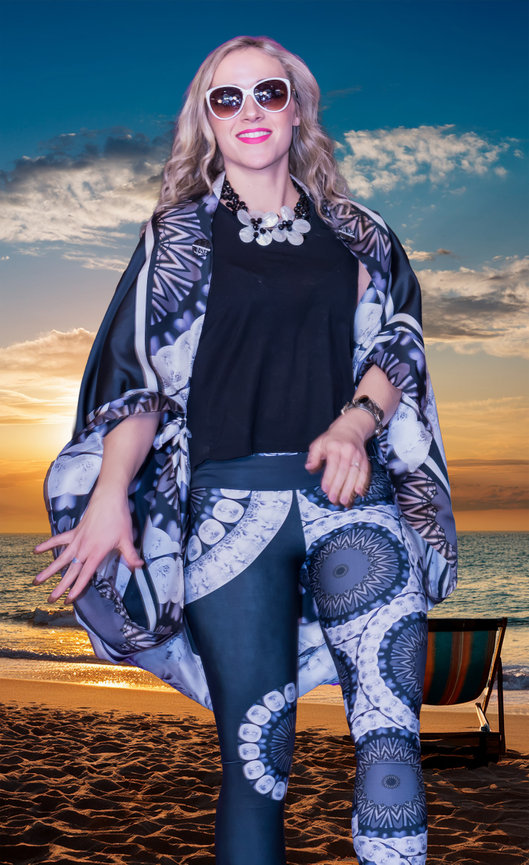 Fashion model on a beach at sunset, wearing white sunglasses, a black top, and a matching black-and-white patterned kimono and leggings with circular mandala designs. Accessories include a floral statement necklace, a bracelet, and a wristwatch.