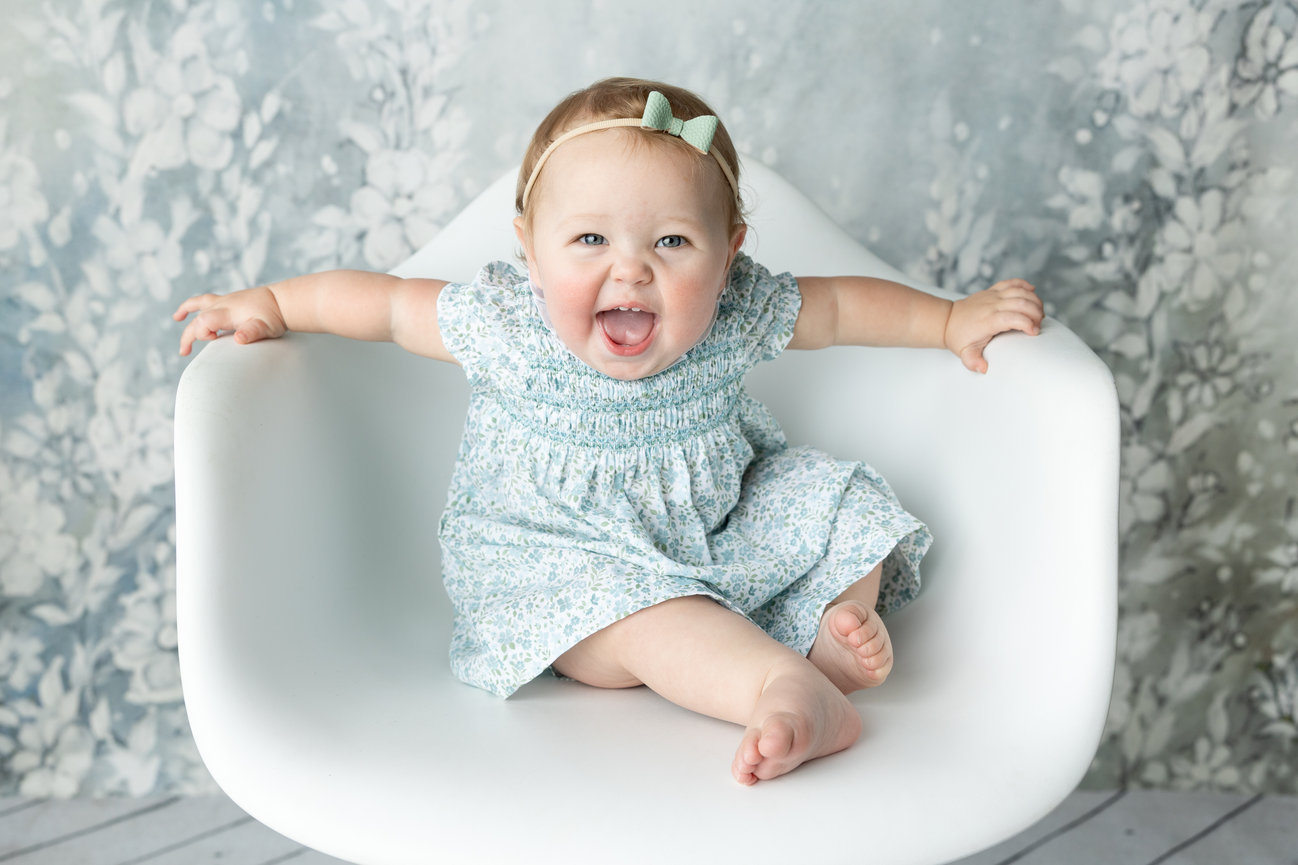 Smiling baby in a floral dress sitting on a white chair against a floral backdrop.