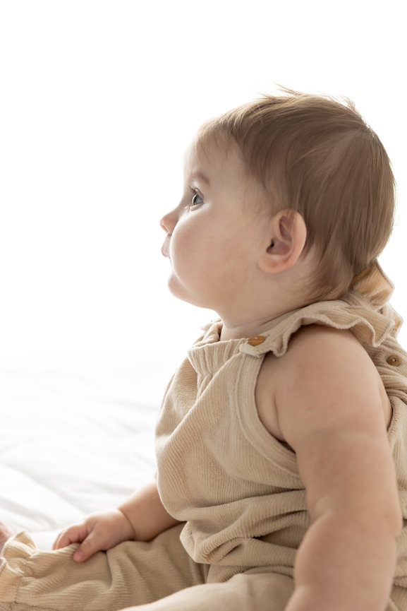 Baby in beige outfit sitting, looking to the side against a white background.