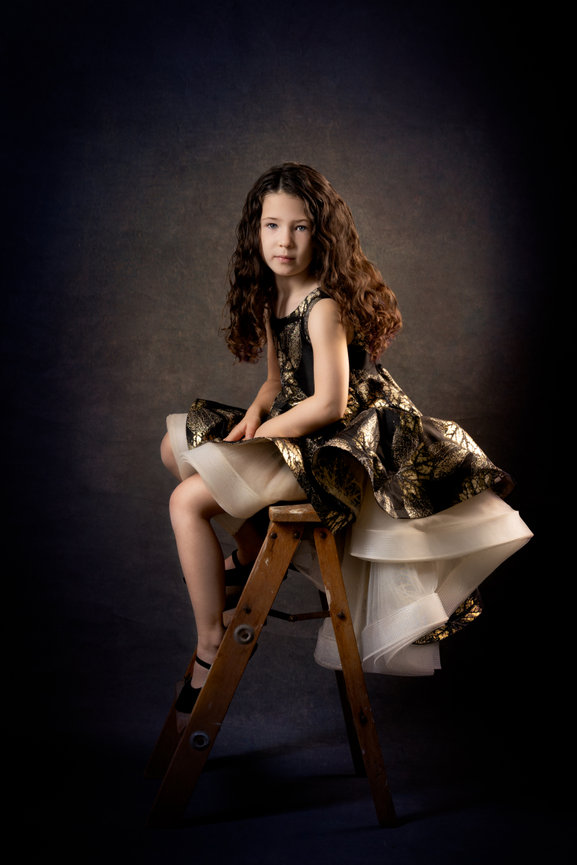 Young girl with long curly hair in a fancy dress sits on a wooden ladder against a dark background.
