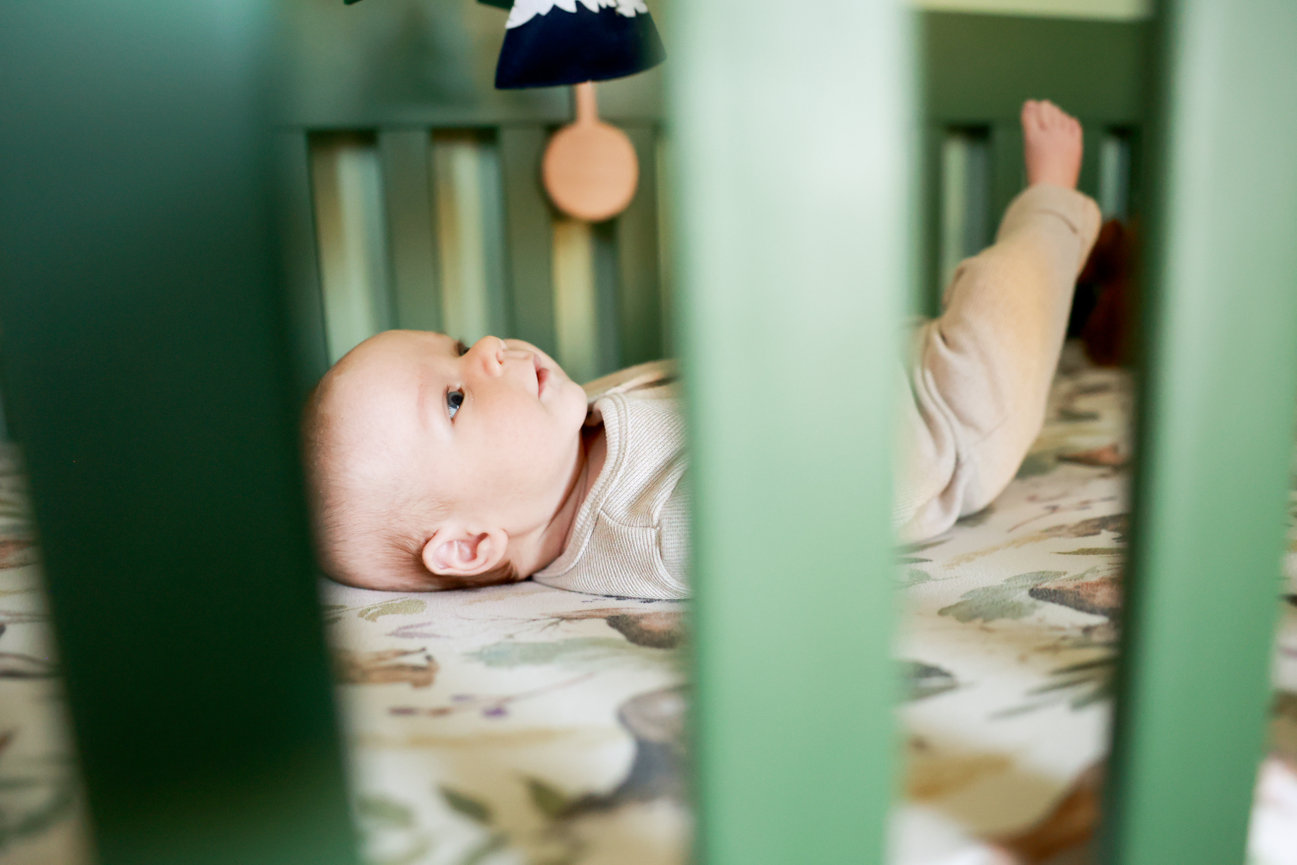 Baby lying in a crib with green bars, wearing a beige outfit, gazing upward.