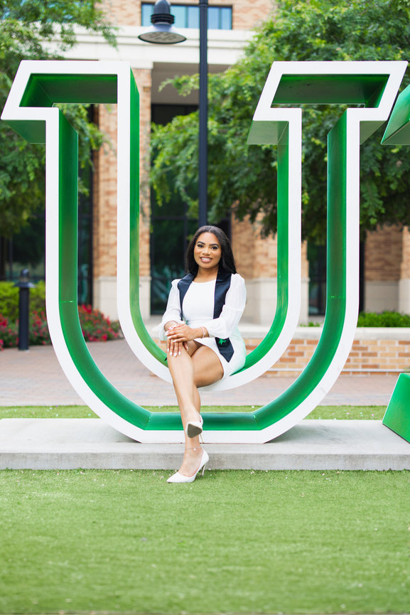 UNT Grad studenton sitting on a large, green letter U sculpture outdoors, with trees and a building in the background.