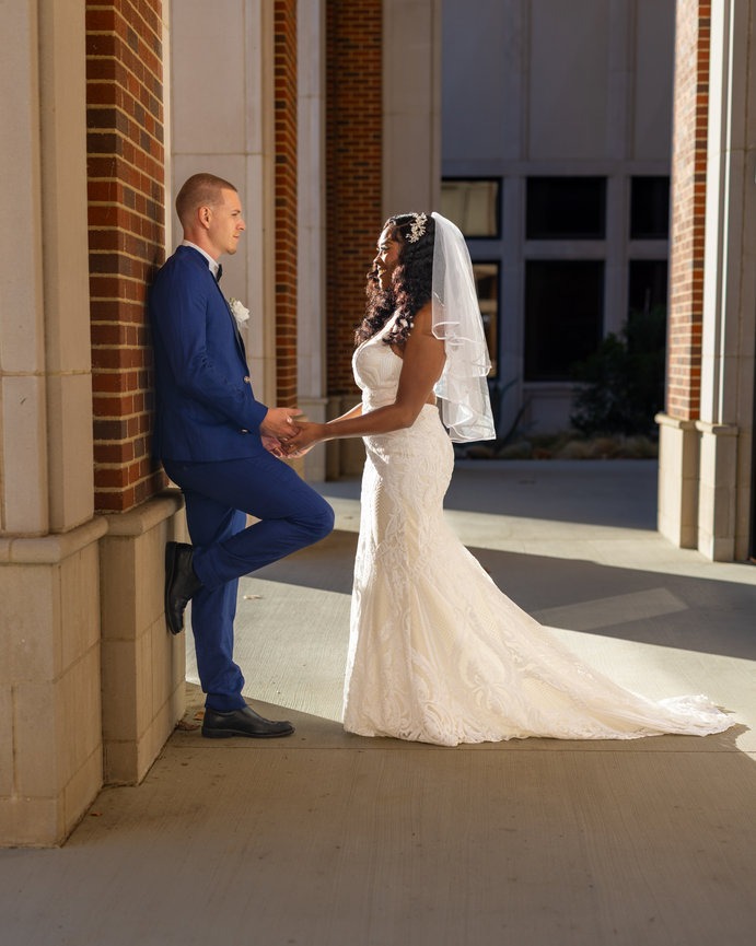 Bride and groom holding hands, facing each other, in a sunlit, brick-walled corridor.