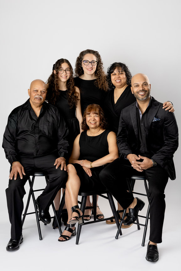 Portrait of six smiling family members in black outfits posed on stools against a white studio background