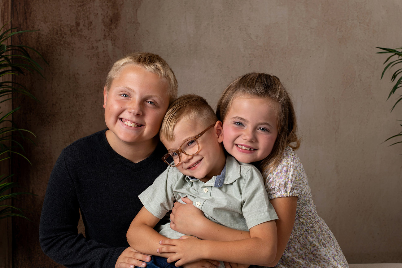 Three smiling children are seated together, with the middle child wearing glasses. The background is neutral with plants. in a studio setting