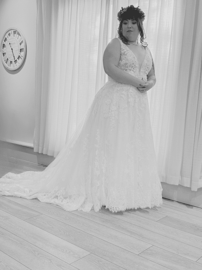 Bride in lace wedding dress with a flower crown, standing by sheer curtains and a wall clock.