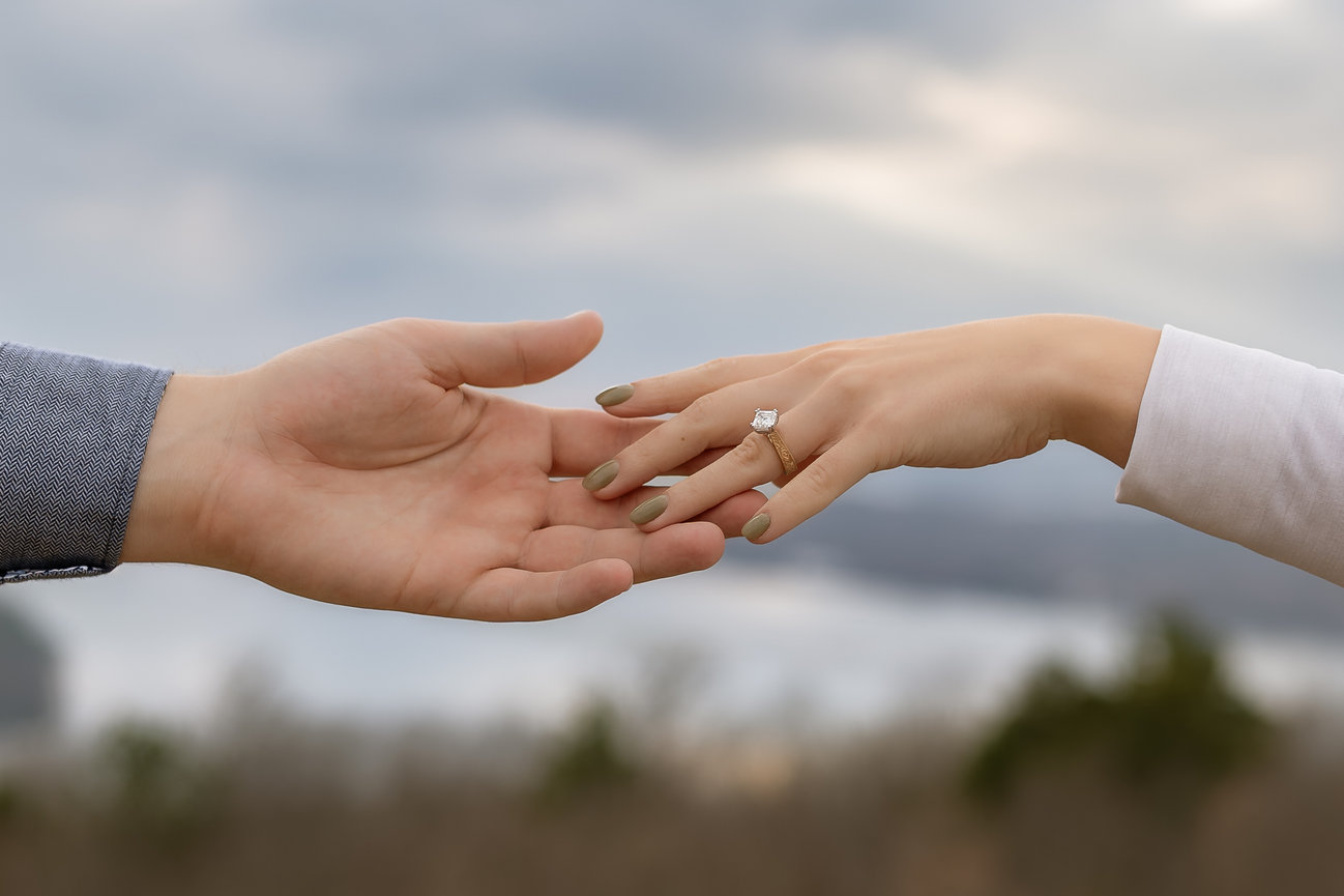 Two hands reaching towards each other outdoors; one with an engagement ring.