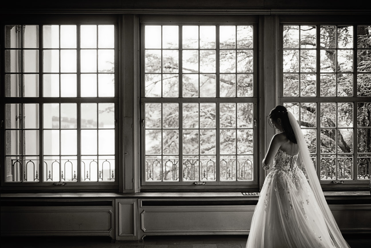 Bride in a white gown standing by large windows in a dimly lit room