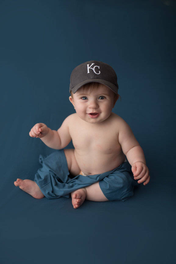 baby boy sitting on royal blue background wear a Kansas City royals hat during baby photoshoot with kansas baby photographer Dreamworks Photography