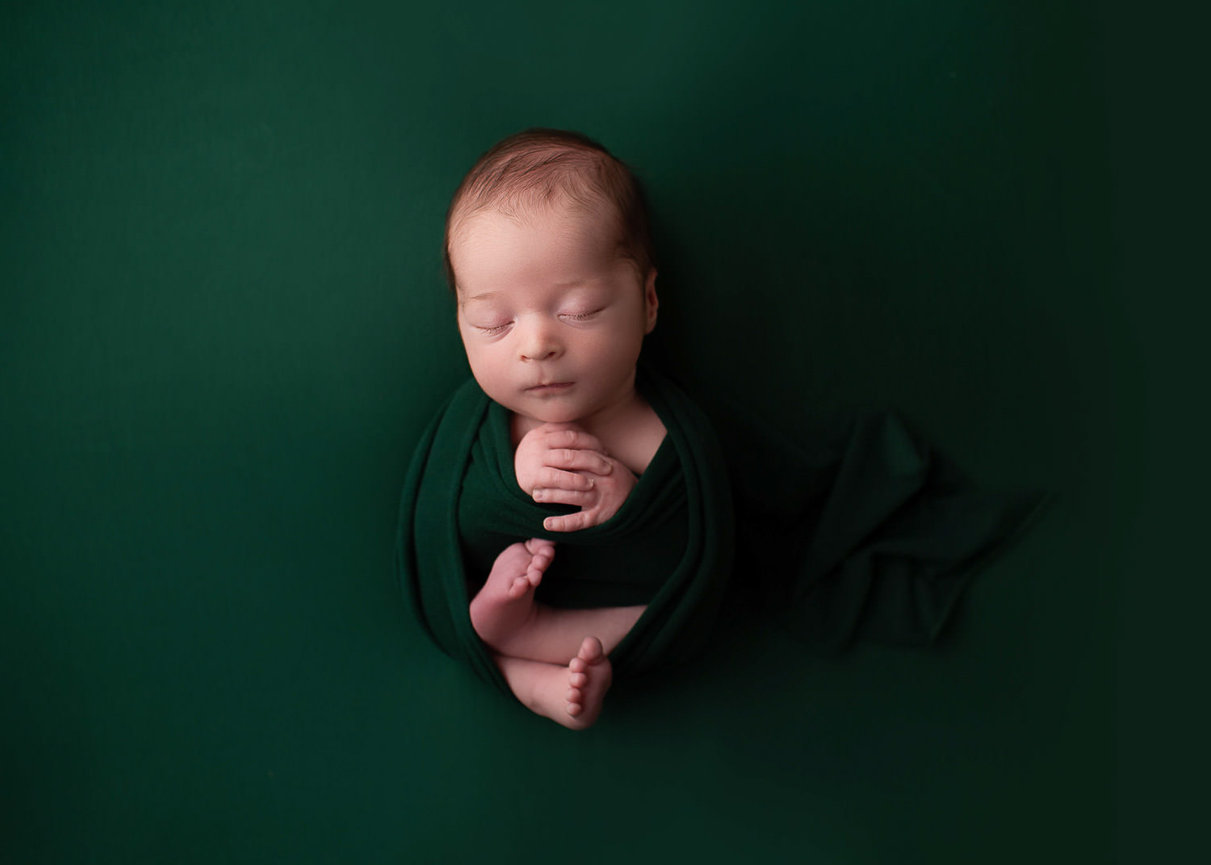 Newborn baby boy laying on hunter green backdrop wrapped in matching fabric during newborn photography session with newborn photographer Dreamworks Photography 