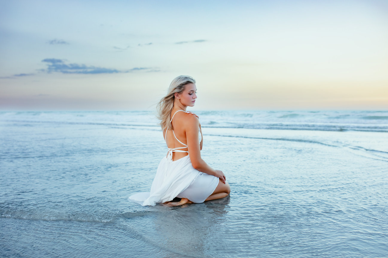 A serene moment during a high school senior photoshoot as a woman in a white dress sits by the shoreline, looking over her shoulder at the tranquil ocean horizon during sunset in jacksonville florida