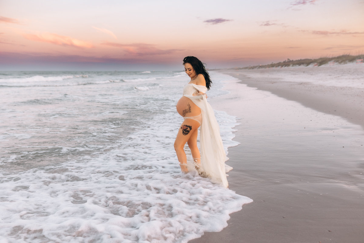 Pregnant woman in a white dress stands at the water's edge on a beach during sunset at Hanna Park Beach during a maternity photography session with Catherine Whitney Photography in Jacksonville Florida