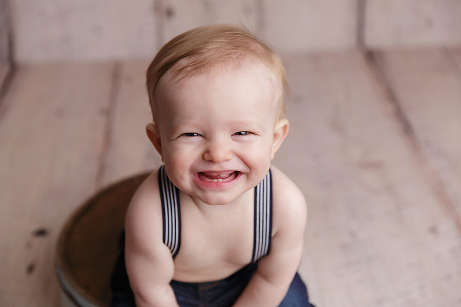 Smiling baby wearing suspenders sitting on wooden floor captured on his first birthday by Omaha, Nebraska Photographer