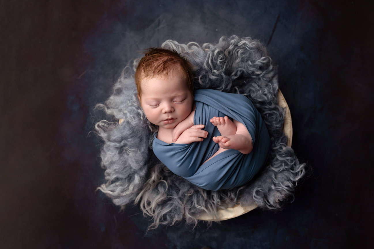 Omaha, Nebraska Newborn Photographer, Sleeping baby wrapped in a blue blanket, lying on a fluffy, gray surface against a dark background.