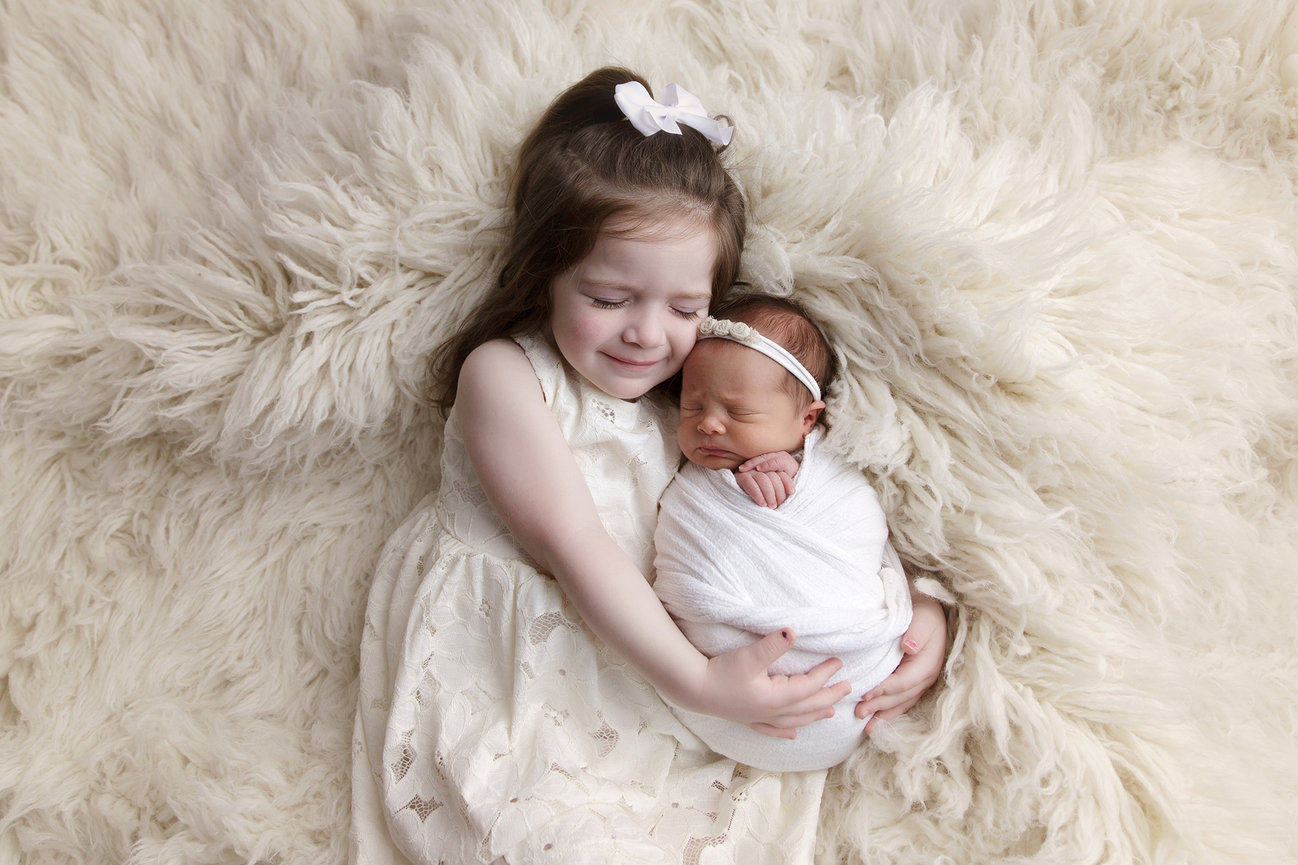 Sisters, Young girl in a white dress cuddles a swaddled newborn baby on a fluffy beige background.