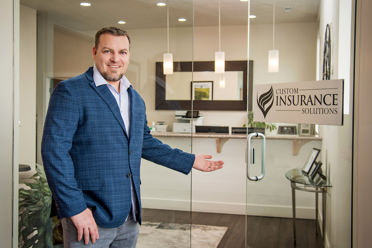 Man in a blue suit standing and gesturing towards an office with a Custom Insurance Solutions sign on the glass door.