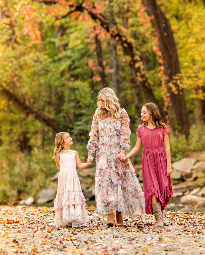 A beautiful mother in a flowing dress looks at her two adorable daughters in pink dresses while walking alongside a creek in the fall in Carmel Indiana.