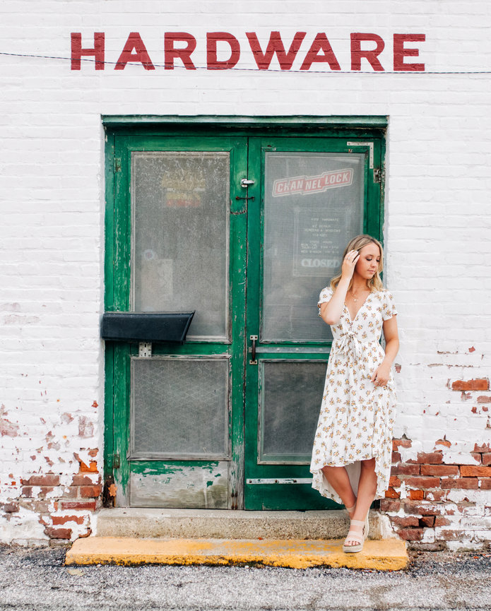 A gorgeous blonde high school senior girl in a flowery dress looking off to the side while standing in front of a hardware store in Noblesville Indiana.