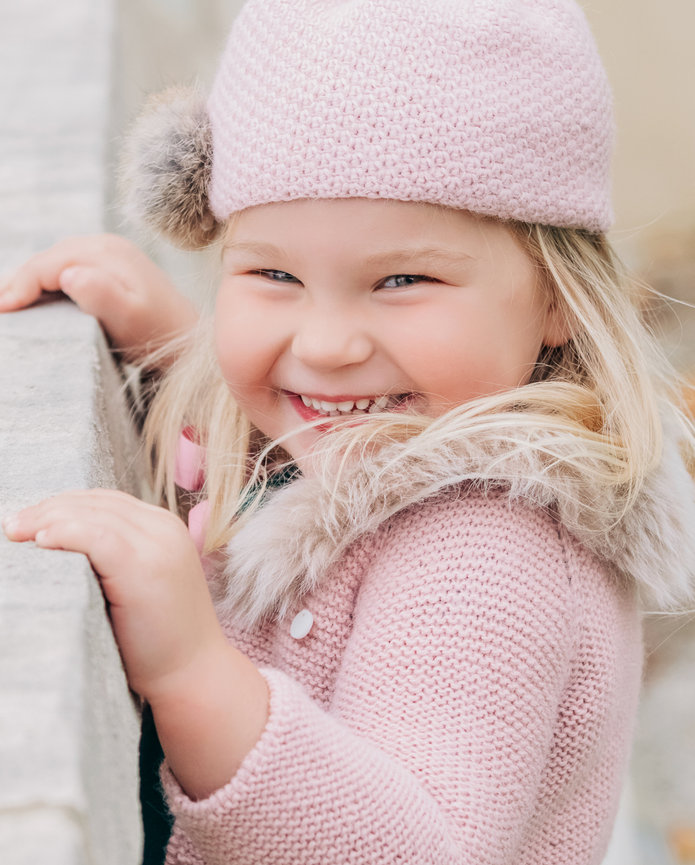 An adorable little girl with a pink knit sweater and matching pink knit stocking hat holds onto a ledge while smiling at the camera in Indianapolis Indiana.