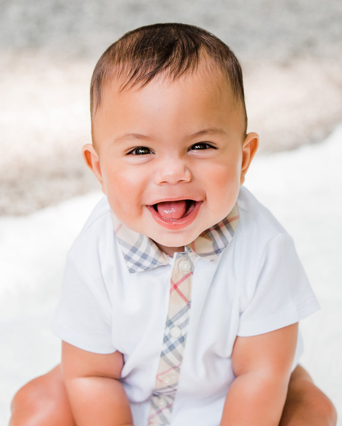 Smiling baby in a white shirt with a patterned tie, sitting on a soft surface outdoors.