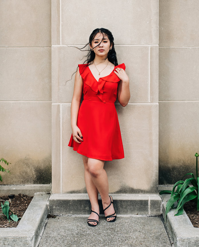 A high school senior in a red dress stands against a stone wall, surrounded by green plants, with a thoughtful expression.