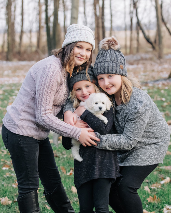 Three people wearing winter clothes and hats, smiling and holding a small white dog in a grassy park with trees in the background.