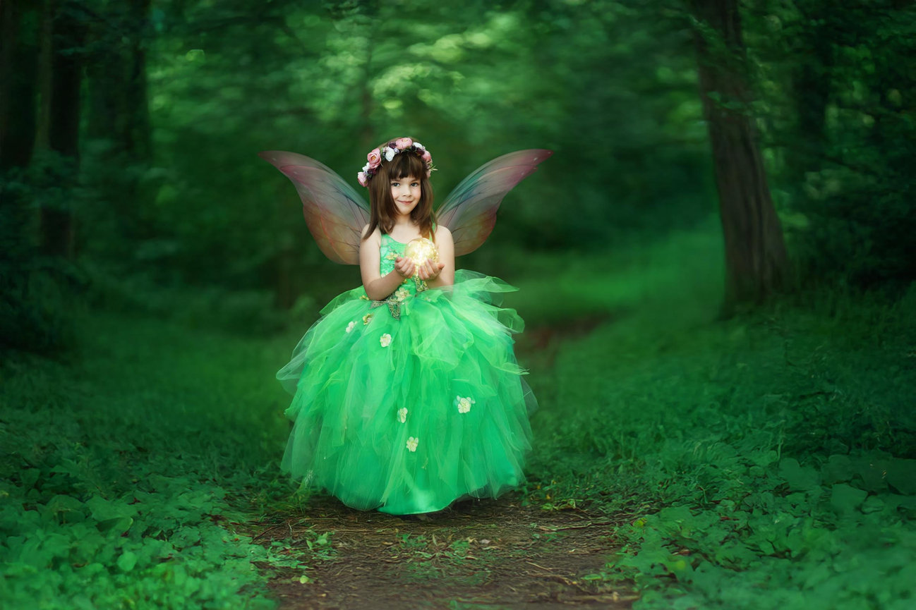 Little girl fairy in a green gown walking through a sunlit meadow during an outdoor fairy session