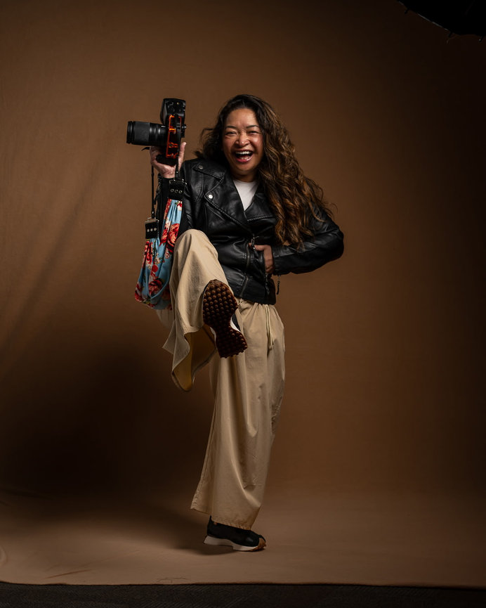 Person in a leather jacket posing energetically with a camera and colorful bag against a brown background as Jo Hayes Images Charlotte Event Portrait Fashion Branding Photographer