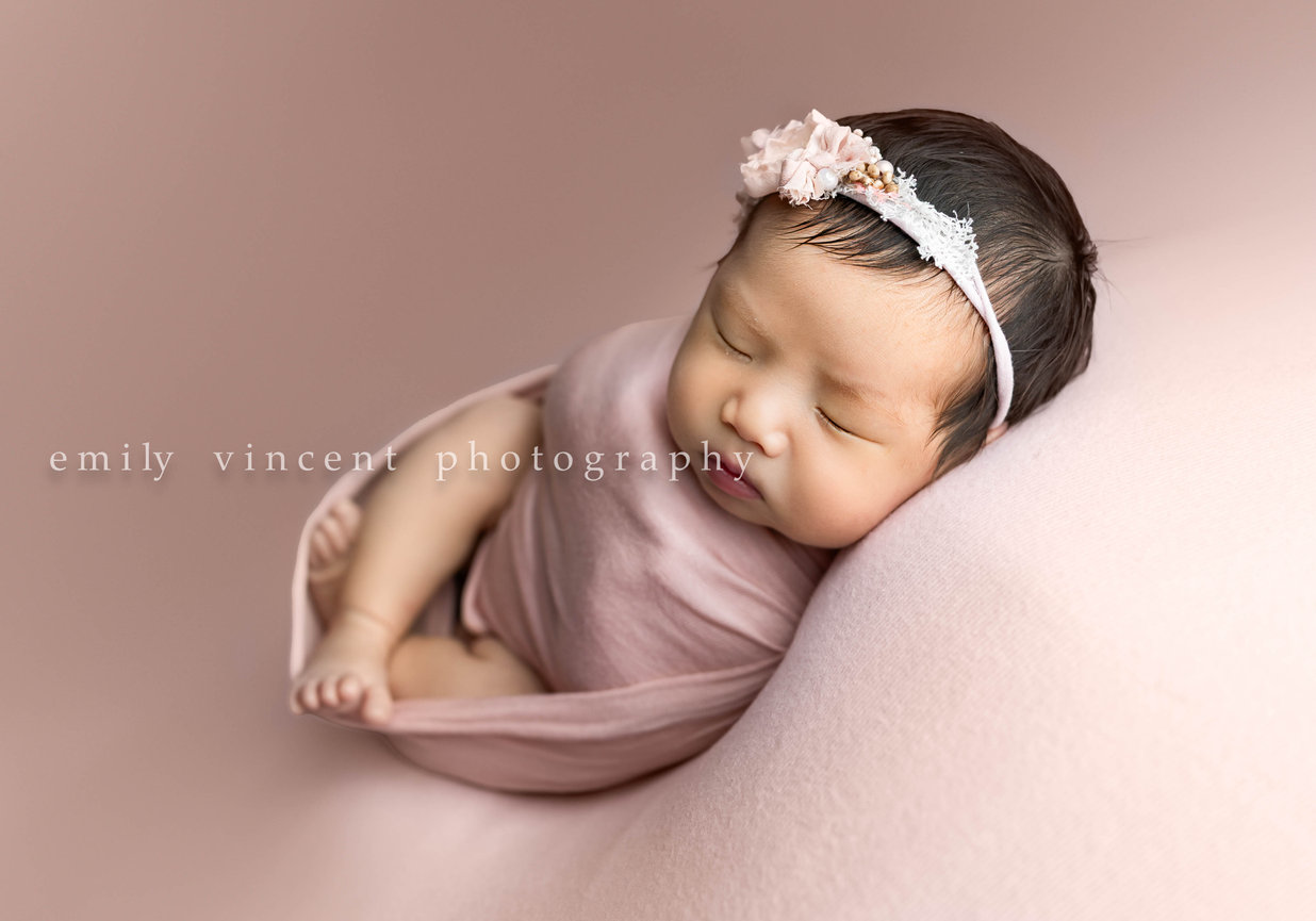 Newborn baby swaddled in white blanket sitting up in wicker chair