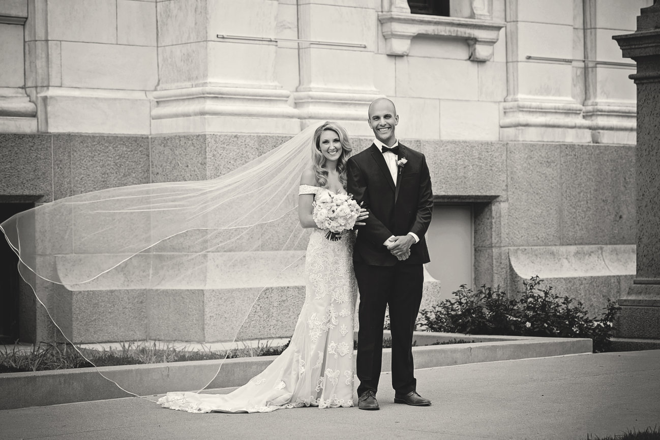 Bride and groom posing outside a stone building, with the bride's veil flowing in the wind.