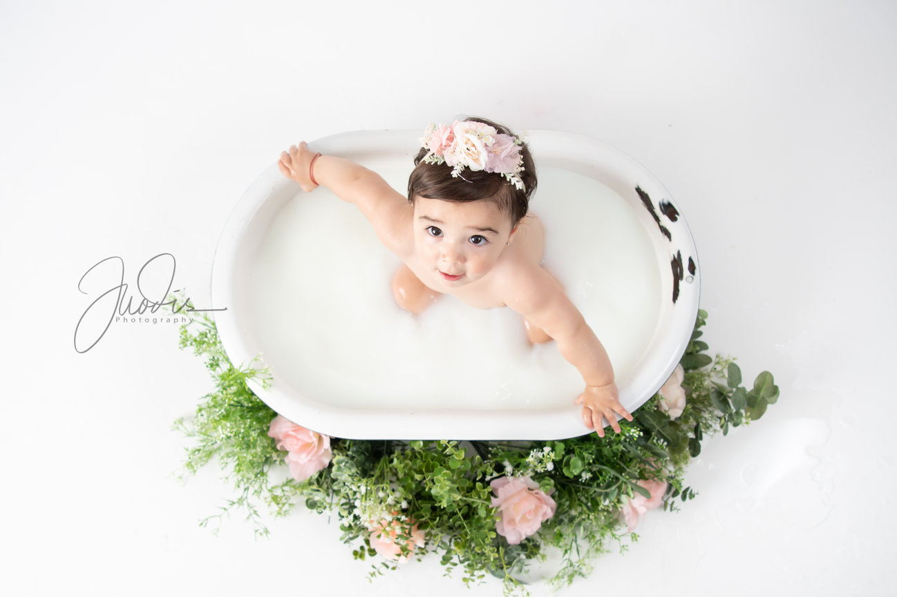 A baby in a vintage bathtub filled with milk, surrounded by flowers and greenery, smiling at the camera.
