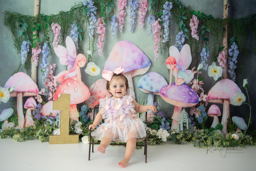 A joyful baby girl in a dress sits on a chair, surrounded by colorful mushroom decorations and greenery.