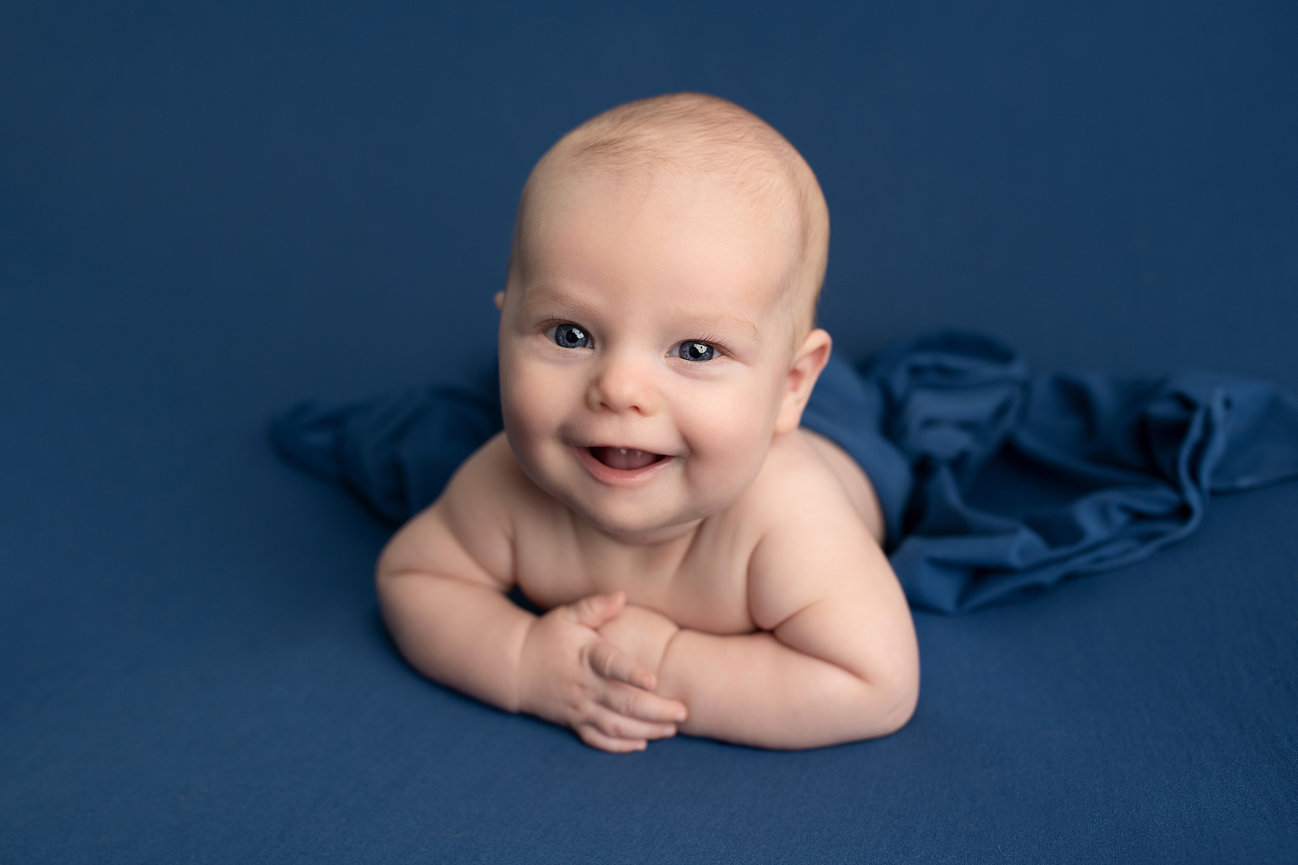 The art of baby photography comes to life as a smiling baby lies on a blue blanket, looking directly at the camera. Propped up on their forearms with a happy expression, the background and blanket in rich, blue is perfect with the babys skin tone.