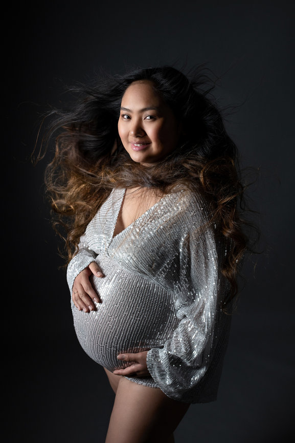 A pregnant woman poses against a dark background, wearing a shimmering, silver, long-sleeved top. Her long, wavy hair is flowing around her. She smiles softly, cradling her belly with both hands.