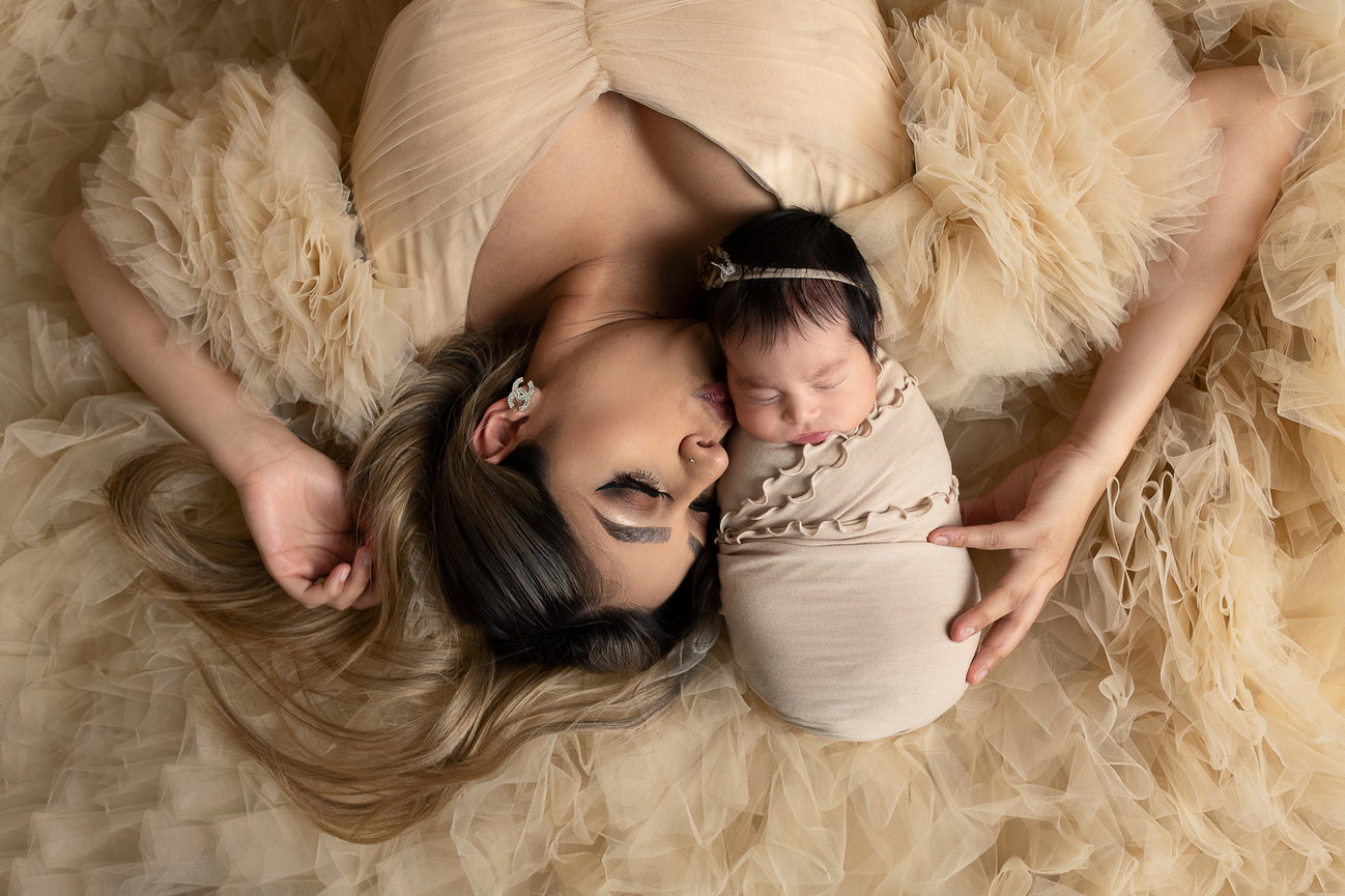 Mother lying on a fluffy, cream-colored surface, eyes closed, gently nestling her swaddled baby wrapped in a matching fabric. Both appear serene and content in their tender embrace, a moment of tranquility of peaceful mornings in Wayne, Illinois.