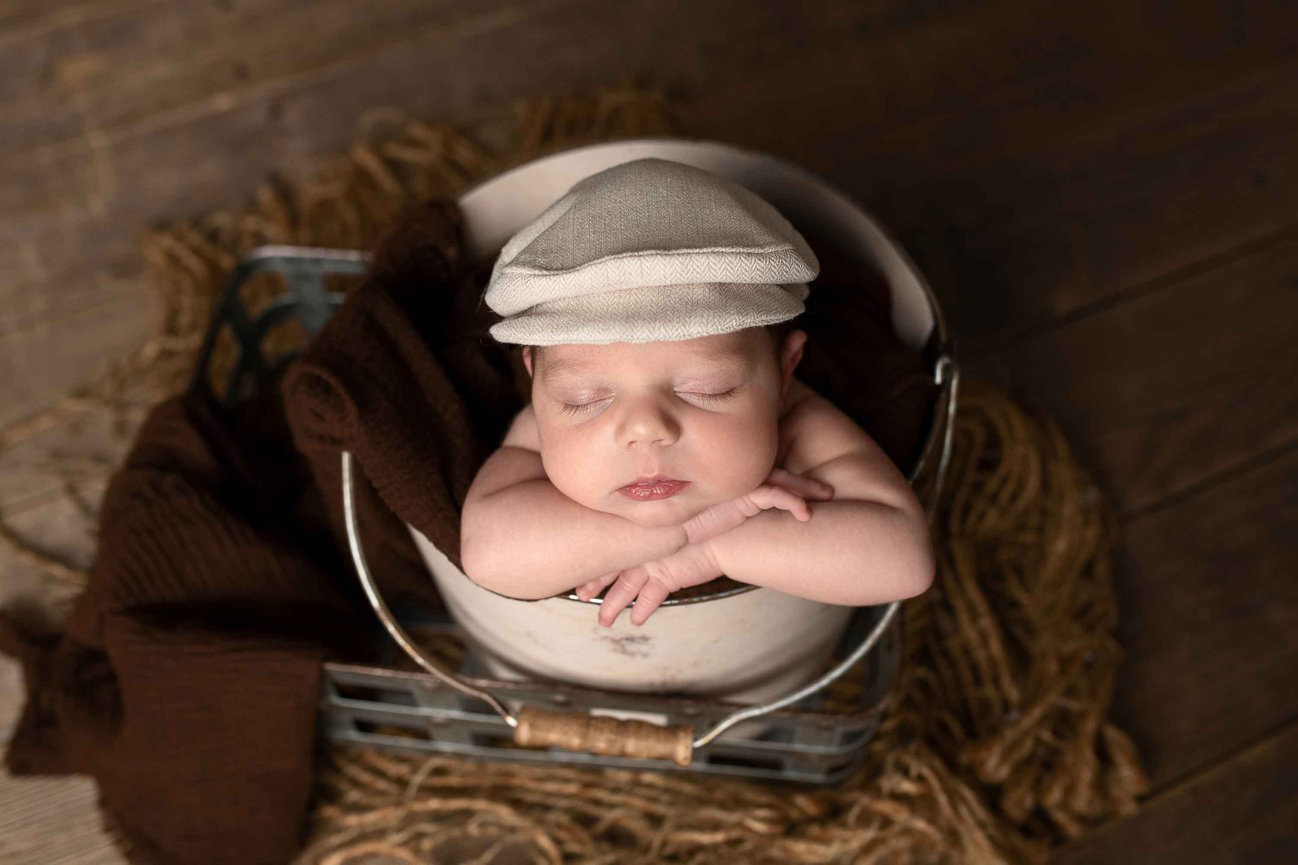 A sleeping baby, wearing a small flat cap, is nestled in a bucket draped with a brown blanket. The setup, accented with subtle Williamsburg Green hues, sits on a wooden floor with burlap touches around the bucket. The babys arms rest on the rim.