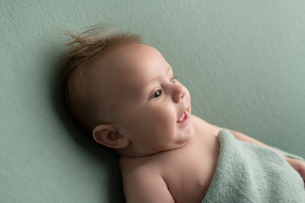 A baby with spiky hair lies on its side, wrapped in a soft green blanket, embodying the essence of baby and maternity photography. The matching green background creates a serene atmosphere as the joyful little one gazes to the side.