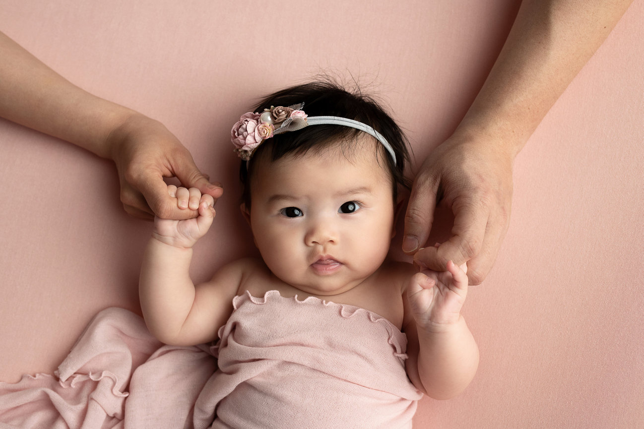A baby wrapped in a pink blanket lies on a soft pink background, creating an image full of warmth and tenderness. The infant, wearing a floral headband, gently clutches the hands of two adults, capturing a heartwarming scene.
